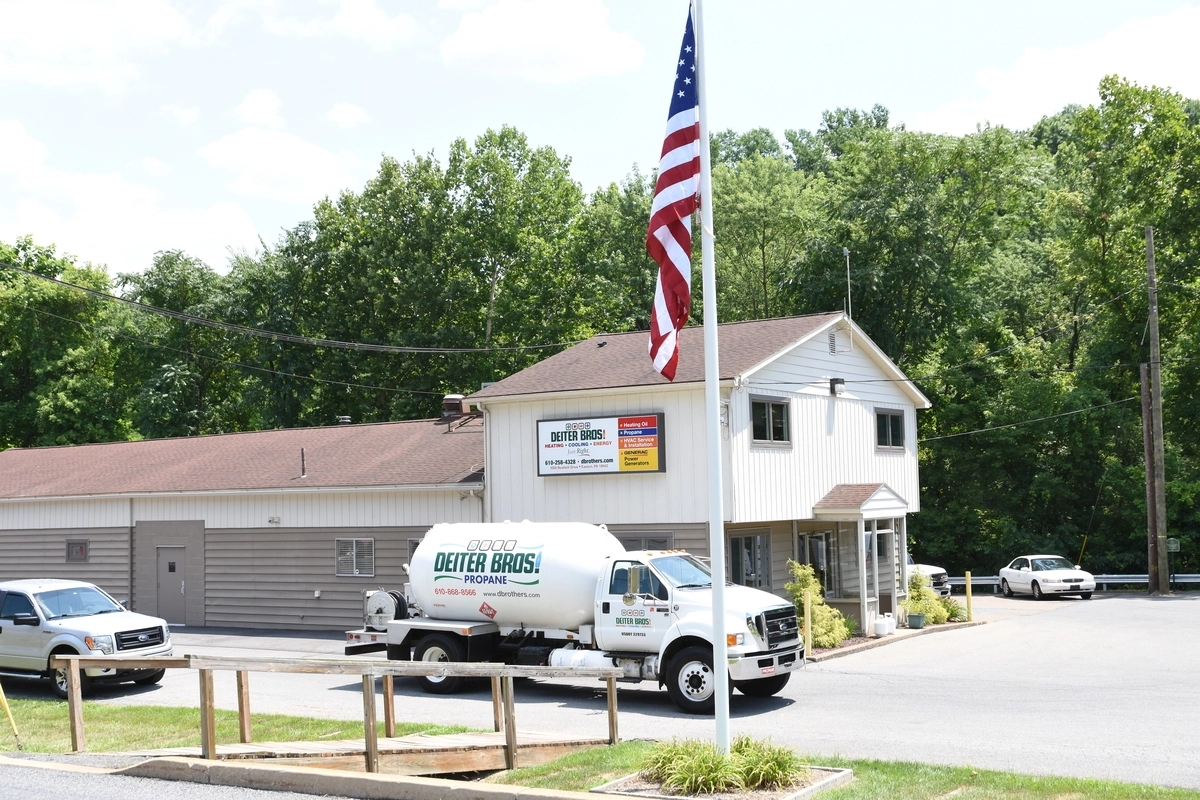 Propane delivery truck parked outside a small HVAC and propane service building with an American flag in the foreground on a sunny day.