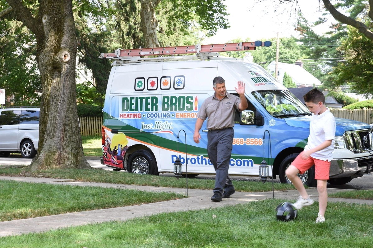 HVAC technician waving to a young boy playing soccer on the lawn, with a branded service van parked on a suburban street in the background.