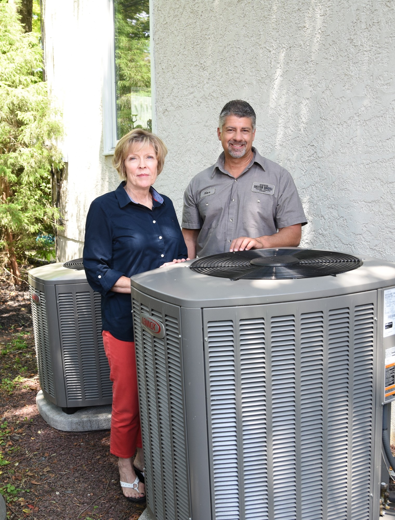 HVAC technician and homeowner standing next to an outdoor air conditioning unit beside a residential home surrounded by greenery.